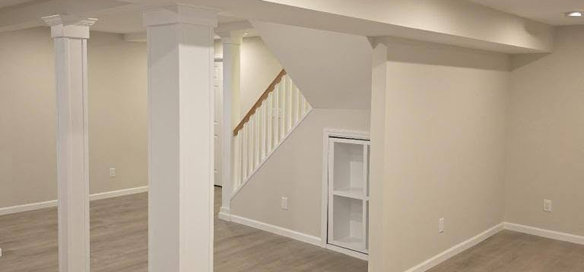 Create Lasting Value and Living Space with These Finished Basement Ideas in Suffolk County Finished basement in Suffolk County with white wrapped support columns, open staircase, built-in shelving and gray vinyl plank flooring